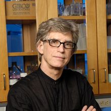 Bespectacled man wearing black shirt with arms folded looks at camera in front of lab cabinet